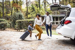 Parents with a toddler riding on a suitcase on wheels, preparing to put luggage into the car boot