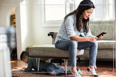 Woman looking at her phone, with child climbing under furniture behind her