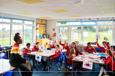 eacher talking to class of primary school children holding up pieces of paper